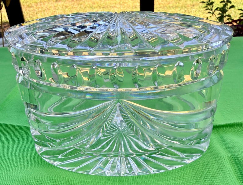 May include: A clear, oval-shaped crystal container with a matching lid. The lid and body feature intricate, geometric patterns, including radial designs and curved lines. The container sits on a green surface, reflecting light and creating a sparkling effect.