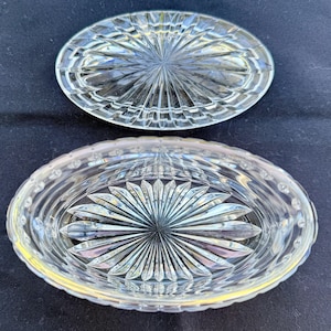 May include: Clear, oval-shaped crystal dish with a lid. The dish and lid feature a starburst design in the center, surrounded by a pattern of vertical and horizontal lines. The dish is on a dark blue surface.