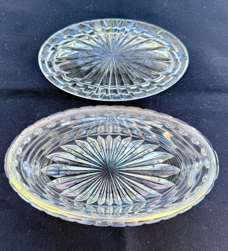 May include: Clear, oval-shaped crystal dish with a lid. The dish and lid feature a starburst design in the center, surrounded by a pattern of vertical and horizontal lines. The dish is on a dark blue surface.