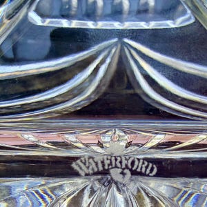 May include: Close-up of a clear crystal object with a geometric design. The word "WATERFORD" is etched into the crystal. The crystal has a reflective quality, with light refracting through the patterned surface.