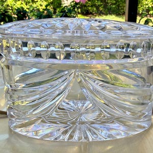 May include: A clear, cut-glass jar with a lid. The jar has a round shape with a decorative pattern of vertical and horizontal cuts. The lid has a starburst design. The jar is likely used for storage or display.