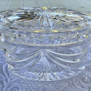 May include: A clear, cut-glass container with a lid. The container is round with a detailed, geometric pattern. The lid has a sunburst design. The glass is thick and reflects light, creating a sparkling effect. The container sits on a white, textured surface.