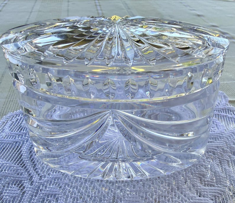 May include: A clear, cut-glass container with a lid. The container is round with a detailed, geometric pattern. The lid has a sunburst design. The glass is thick and reflects light, creating a sparkling effect. The container sits on a white, textured surface.