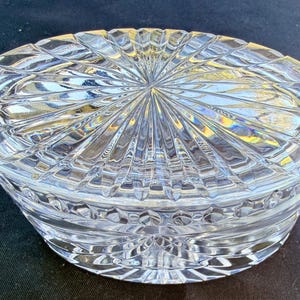 May include: A clear, oval-shaped crystal trinket box with a lid. The lid and base feature a detailed, geometric pattern of radiating lines and diamond shapes. The box is designed to catch and reflect light, creating a sparkling effect.