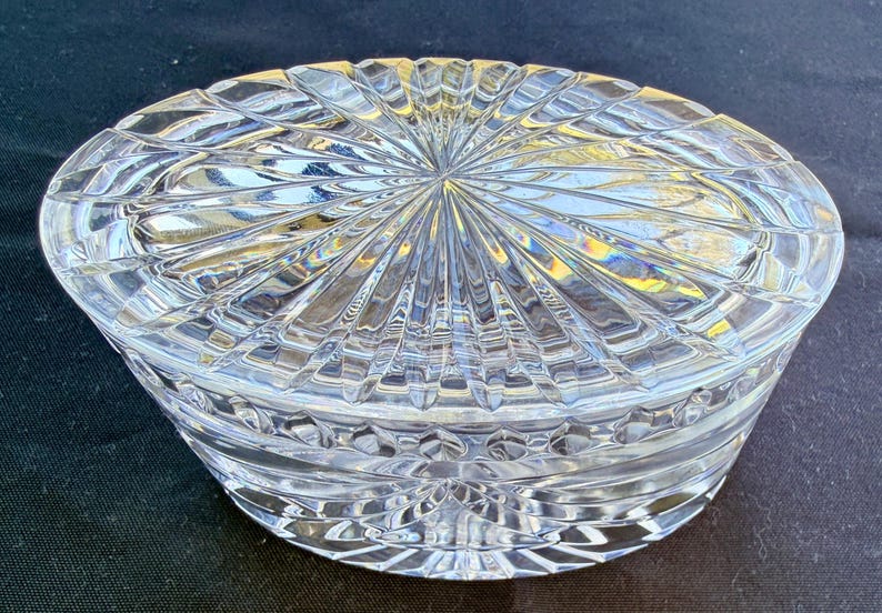 May include: A clear, oval-shaped crystal trinket box with a lid. The lid and base feature a detailed, geometric pattern of radiating lines and diamond shapes. The box is designed to catch and reflect light, creating a sparkling effect.