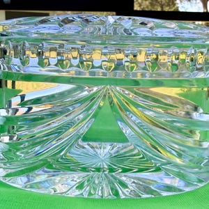 May include: A clear, cut-glass jar with a lid. The jar has a rectangular shape with intricate geometric patterns, including vertical ridges and swirling designs. The lid also features a detailed, textured surface. The jar is likely used for storage or display.