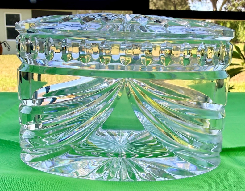 May include: A clear, cut-glass jar with a lid. The jar has a rectangular shape with intricate geometric patterns, including vertical ridges and swirling designs. The lid also features a detailed, textured surface. The jar is likely used for storage or display.
