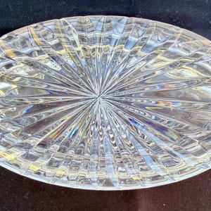 May include: Oval-shaped, clear crystal dish with a starburst design emanating from the center. The crystal dish has a textured surface with a repeating pattern of lines and angles, creating a sparkling effect. The dish is likely used for decorative purposes.