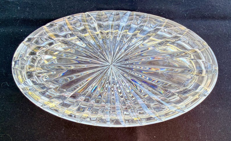 May include: Oval-shaped, clear crystal dish with a starburst design emanating from the center. The crystal dish has a textured surface with a repeating pattern of lines and angles, creating a sparkling effect. The dish is likely used for decorative purposes.