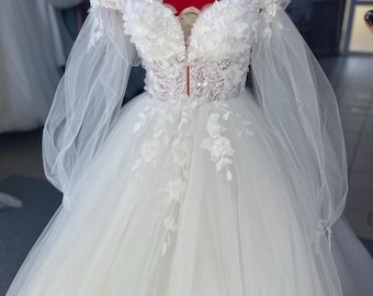 May include: A white off-the-shoulder wedding dress with long sleeves and a tulle skirt. The bodice is adorned with delicate lace and floral appliques.