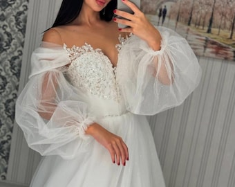 May include: A white tulle wedding dress with a sweetheart neckline and off-the-shoulder straps. The dress has a fitted bodice with beaded lace detailing and a flowing skirt. The sleeves are long and puffy, made of sheer tulle.
