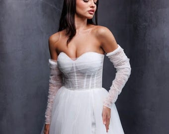 May include: A strapless white wedding dress with a sweetheart neckline and a sheer, structured bodice. The dress features a full, flowing skirt and matching sheer, long sleeves. The gown is designed with a romantic and elegant aesthetic.