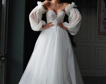 May include: A white off-the-shoulder wedding dress with a fitted bodice and a full tulle skirt. The dress has long, puffy sleeves with lace detailing and a sweetheart neckline.