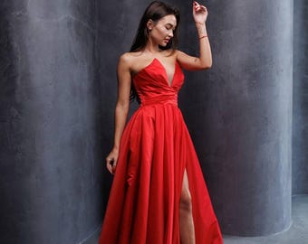 May include: A woman wearing a red strapless gown with a thigh-high slit. The gown has a fitted bodice and a flowing skirt. The woman is standing in front of a gray background.