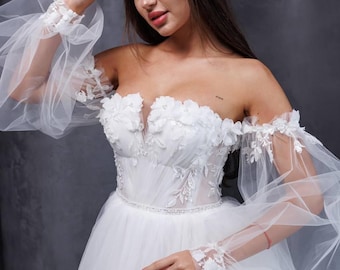 May include: A white off-the-shoulder wedding dress with a fitted bodice and a full skirt. The dress features delicate floral lace appliques and a beaded belt. The sleeves are long and puffy, made of tulle.