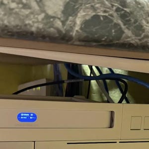 May include: A cream-coloured electronic device with a blue display and buttons is mounted in a rack. Blue and black cables are connected to the device. The device is installed under a countertop with a marble pattern.