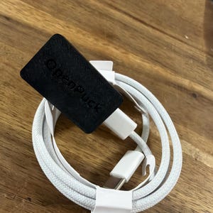 May include: A white braided charging cable with a USB-C connector and a black rectangular attachment featuring the embossed text OpenPuck. The cable is coiled and secured with a white paper band, showing a standard connector at one end.