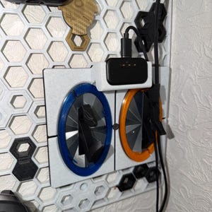 Peut inclure: Un support mural à grille hexagonale blanche doté de deux stations de charge circulaires sur le thème des portails, l'une avec un anneau bleu et l'autre avec un anneau orange. Un chargeur noir est fixé au-dessus, avec un câble noir.