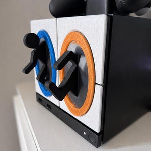 Könnte beinhalten: Ein schwarzer Controller-Ständer mit zwei weißen quadratischen Paneelen und blau-orangen kreisförmigen Portalringen. Zwei schwarze Strichmännchen sind an den Portalen befestigt, eines kommt aus dem blauen Ring und eines geht in den orangen Ring.