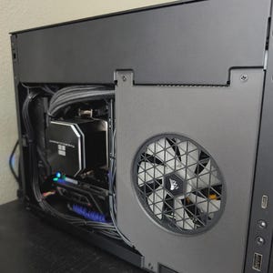 Peut inclure: Un boîtier d'ordinateur noir avec des composants internes visibles. Un grand ventilateur avec un motif géométrique est sur le côté. Le boîtier possède un refroidisseur à eau Thermaltake et divers câbles. Le boîtier est sur une surface sombre.