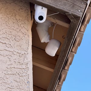 Puede incluir: Un sistema de cámara de seguridad blanco está montado debajo de un alero de madera. El sistema incluye una cámara con lente y un componente separado y redondeado, todo conectado por cableado blanco. La cámara está posicionada para monitorear un área exterior.