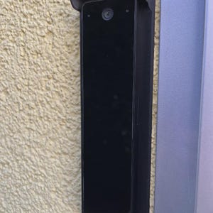 May include: A black rectangular doorbell camera fixed to a textured, light-yellow wall. The camera has a lens and is in a black frame with a small, curved hood. A vertical, grey panel is to the right.