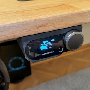 May include: Black SteelSeries audio controller mounted under a wooden desk. The controller has a volume dial, a small screen displaying volume level, and the SteelSeries logo. The screen shows the word "VOLUME" and a battery indicator.