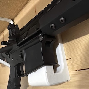 May include: A black rifle with a white plastic mount attached to a wooden wall. The rifle has a black handguard with a few screws visible.