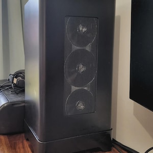 Puede incluir: Una torre de ordenador negra con forma rectangular y un panel frontal de malla con tres aberturas circulares para ventilador. La torre se asienta sobre una base ligeramente elevada y tiene esquinas redondeadas.