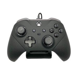 May include: A black Xbox controller with a textured grip and a black stand. The controller has a black and silver design with a black Xbox button.