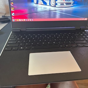 Puede incluir: Un ordenador portátil negro con un teclado completo y un panel táctil plateado. La pantalla muestra una imagen colorida de un coche de carreras. El portátil está abierto y listo para usar.