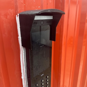 May include: A black intercom system with a keypad and screen is mounted on a red corrugated metal wall. The intercom has a protective hood and a camera. The screen displays the text "Enter PIN".