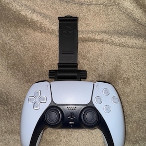 May include: A white PlayStation 5 DualSense controller with a black phone holder attached to the top. The controller has black buttons and a black PlayStation logo.
