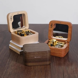 May include: Three wooden music boxes with hinged lids and internal mirrors. Two boxes are open, revealing gold-coloured mechanical movements, while one is closed. Each lid features a small treble clef symbol etched into the light or dark wood.