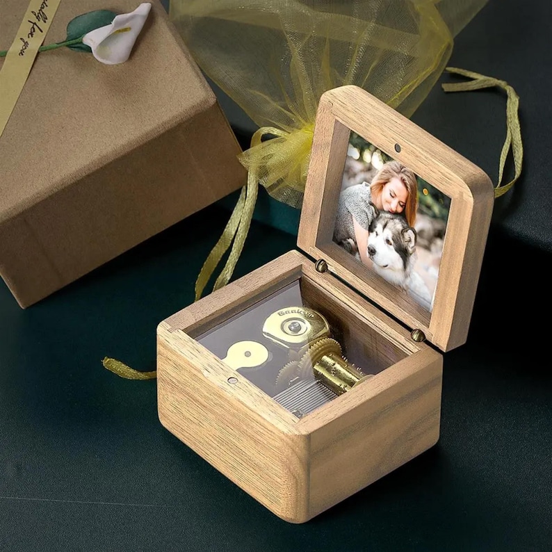 May include: A wooden music box with a clear lid, revealing a gold-colored music mechanism. The box has a hinged lid with a photo frame insert, showing a woman holding a dog. The box is sitting on a green surface.