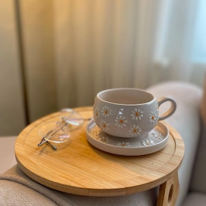 May include: A white ceramic mug and matching saucer set featuring a raised daisy pattern with yellow centers, accompanied by a pair of clear-framed eyeglasses resting on a round wooden tray designed to fit over an armrest.
