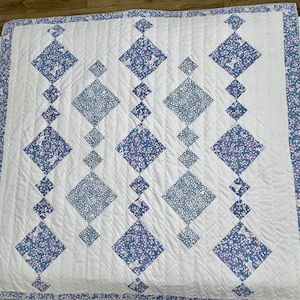 May include: A white quilt with a diamond pattern. The diamonds are made of a blue and pink floral fabric. The quilt is quilted with a white thread.