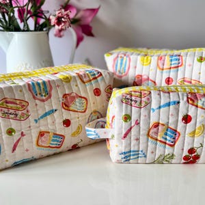 May include: Three rectangular quilted pouches featuring a white fabric base with a repeating pattern of sardine tins, individual sardines, lemon slices, tomatoes, and olives. Each pouch has a yellow and white checkered zipper and trim.