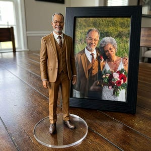 May include: A 3D-printed figurine of a man in a brown suit, standing on a clear acrylic base, next to a framed photograph of the same man with a woman. The photo is in a black frame. The figurine and photo are on a wooden table.