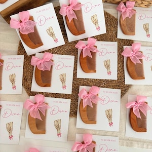 Può includere: Una collezione di pettini in legno fissati su cartoncini bianchi, ognuno decorato con un fiocco di nastro rosa, un'illustrazione di due mani che si tengono e il testo "TU BI XER HATI" e "Duaa" stampato in corsivo sul cartoncino.
