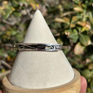 May include: A silver bangle bracelet with a black and white inlay design. The bracelet is displayed on a white cone-shaped stand with a wooden base.