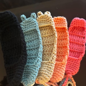 Op de afbeelding: Een set van vijf gehaakte items in zwart, lichtblauw, beige, oranje en roze. Elk stuk heeft een gestructureerd, geribbeld steekpatroon met twee kleine afgeronde uitsteeksels aan de bovenrand en lange bijpassende garenlinten aan de onderkant.