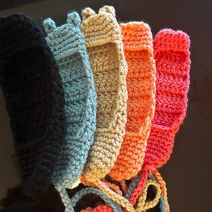 Op de afbeelding: Vijf gehaakte items in zwart, lichtblauw, beige, oranje en roze. Elk stuk heeft een gestructureerd, geribbeld steekpatroon met een kleine opening aan de bovenkant en lange, bijpassende gehaakte koorden die aan de onderkant van elk item hangen.
