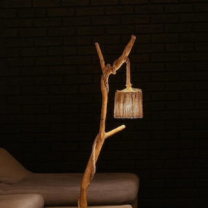 May include: A rustic floor lamp crafted from a natural tree branch, featuring a woven lampshade. The lamp has a wooden base and a rope detail. The warm light illuminates a dark room with a sofa and a striped rug.