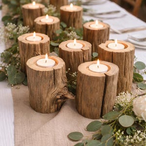 May include: A table setting with wooden candle holders, each holding a lit candle. The holders are made from tree trunk sections, arranged on a beige runner with greenery and white flowers. The candles are small, round, and white, creating a warm ambiance.
