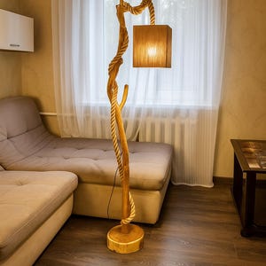 Op de afbeelding: Een houten vloerlamp met een natuurlijk, rustiek ontwerp. De lamp heeft een gedraaid touwdetail en een vierkante lampenkap. De basis is een ronde houten schijf. De lamp staat in een woonkamer.