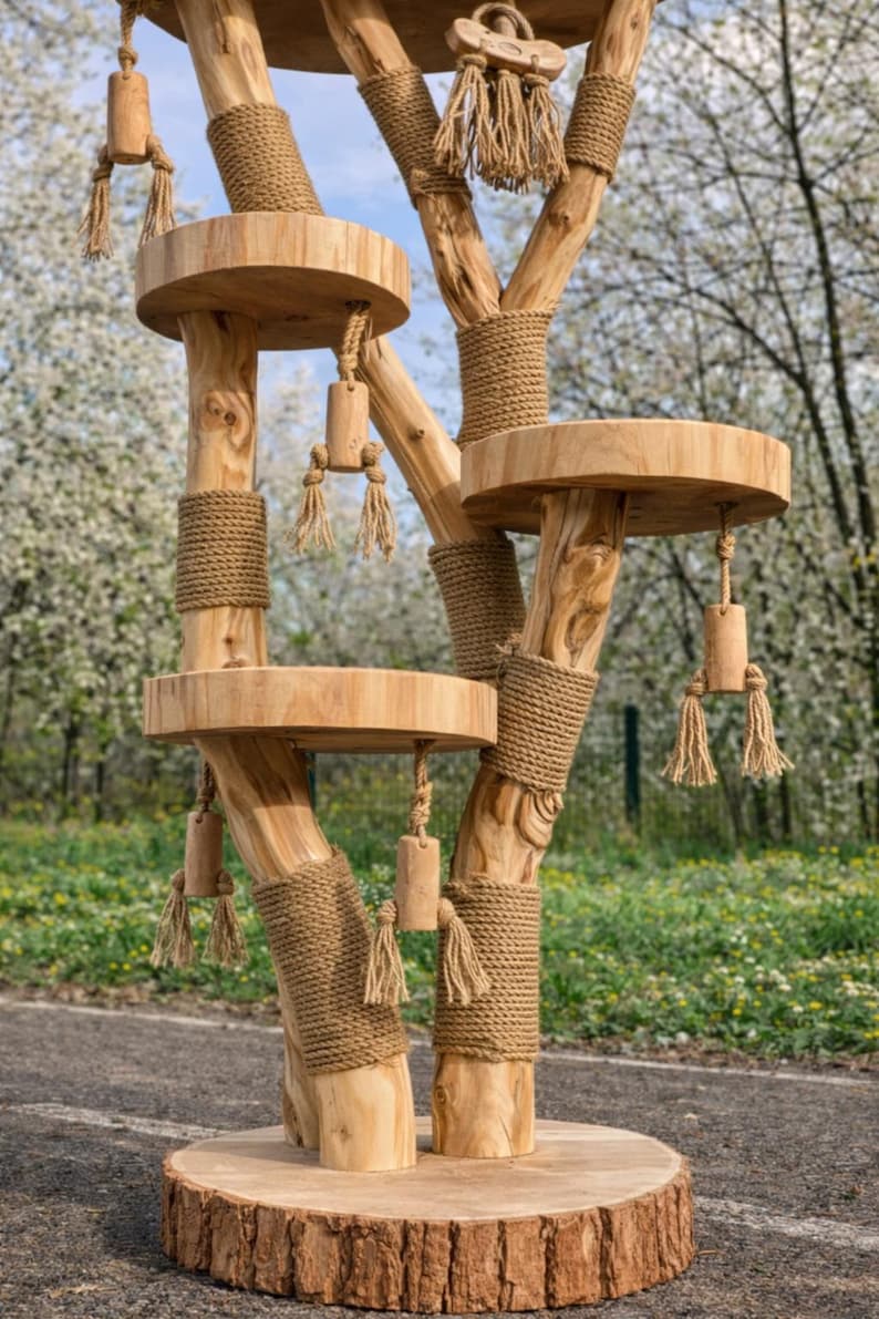 May include: A natural wood cat tree with multiple platforms and scratching posts. The tree features round platforms, rope-wrapped supports, and dangling toys. The base is a large, circular wooden slab. The background shows a blurred green lawn and white flowering trees.