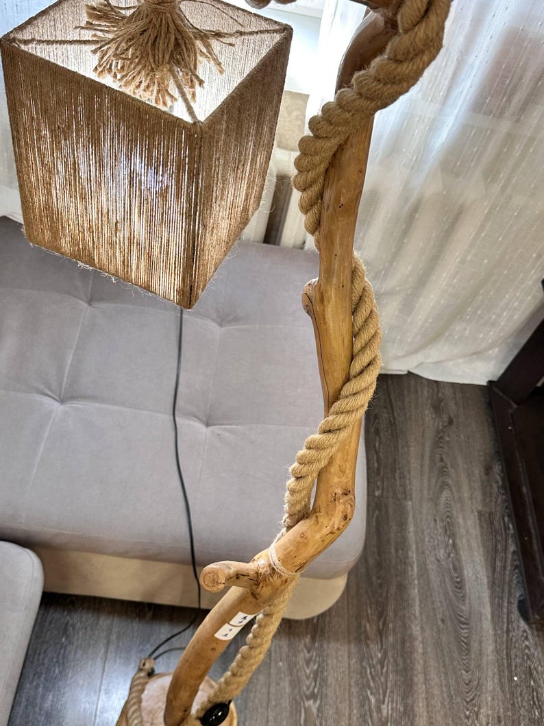 Op de afbeelding: Een vloerlamp met een natuurlijke houten frame en een vierkante lampenkap. De lampenkap is gemaakt van geweven bruin materiaal. Een dik, bruin touw is om de houten paal gewikkeld. De lamp is ingeschakeld en het licht schijnt door de lampenkap.