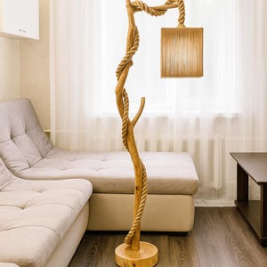 Op de afbeelding: Een unieke vloerlamp gemaakt van natuurlijk hout en touw. De lamp heeft een boomachtige structuur met een vierkante lampenkap. Het touw is om de houten paal gewikkeld, wat een rustieke uitstraling geeft. De lamp staat op een ronde houten voet.