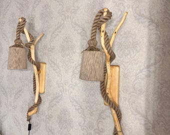 May include: Two wall-mounted lamps featuring natural light-colored wooden branches as the main structure. Each lamp has a thick, tan-colored rope wrapped around the branch, supporting a cylindrical, textured tan lampshade and a black power cord.